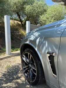 BMW X5 M gris clair avec jantes sport noires et freins bleus, garée sur gravier sous les arbres. Logo "leboncoin".