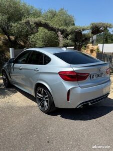 BMW X6 argent vue arrière garée, plaque 'A7 CONCEPT'. Jantes sportives, double échappement. Lumière du jour, arbres anciens en fond.