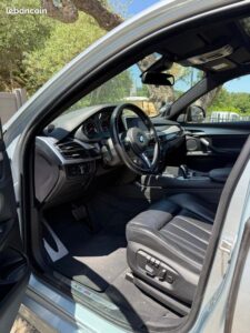 Intérieur détaillé d'un BMW X6 M gris: tableau de bord, volant en cuir, siège conducteur noir et logo X6 M au sol.