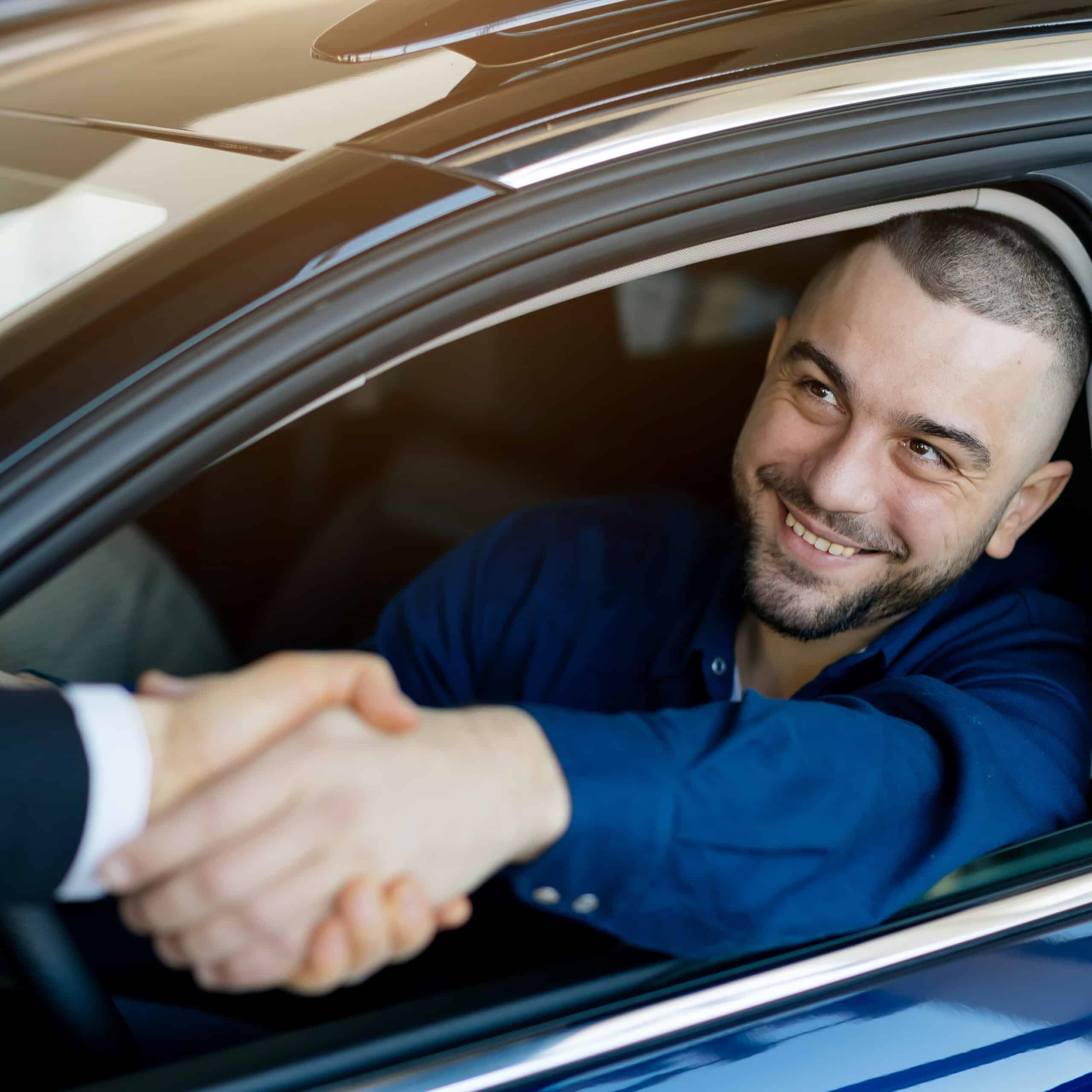 Homme souriant dans une voiture bleue, serrant la main d'une personne en costume. Scène d'achat ou vente de voiture, client satisfait.