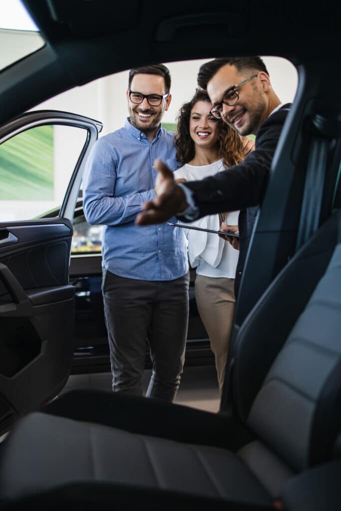 Agent auto souriant présente une nouvelle voiture à un couple heureux dans un concessionnaire moderne.