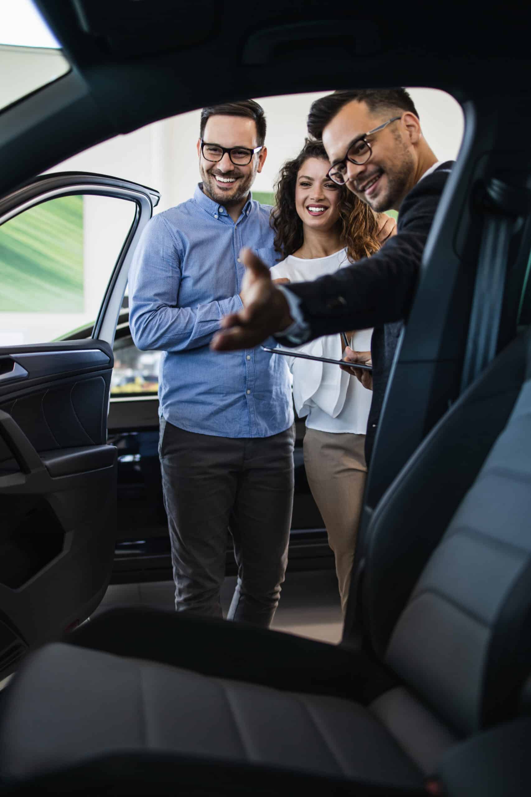 Agent auto souriant présente une nouvelle voiture à un couple heureux dans un concessionnaire moderne.