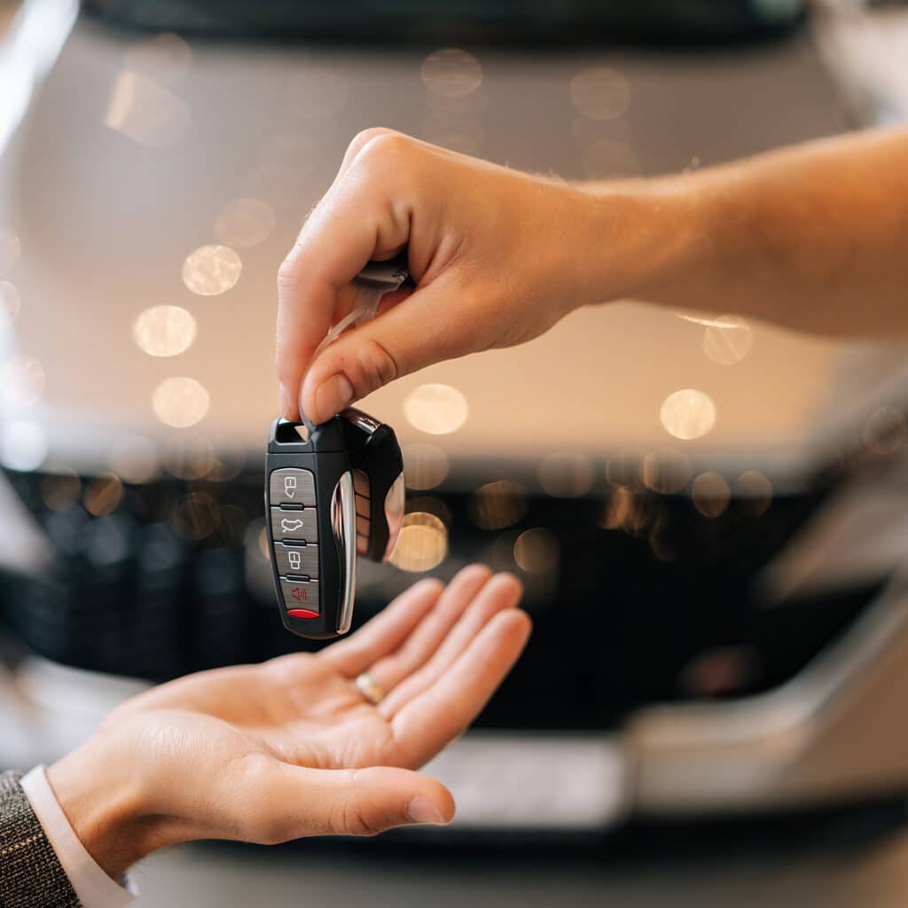 Clé de voiture remise: Votre nouvel achat auto Remise d'une clé de voiture moderne noire et argentée d'une main à l'autre, devant un véhicule neuf flou.