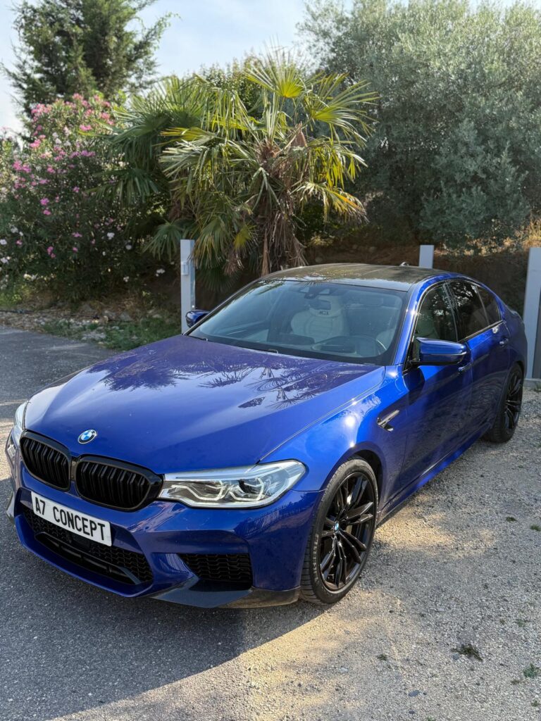 BMW M5 bleue vif au soleil, avec calandre noire et plaque "A7 CONCEPT", entourée de verdure méditerranéenne.