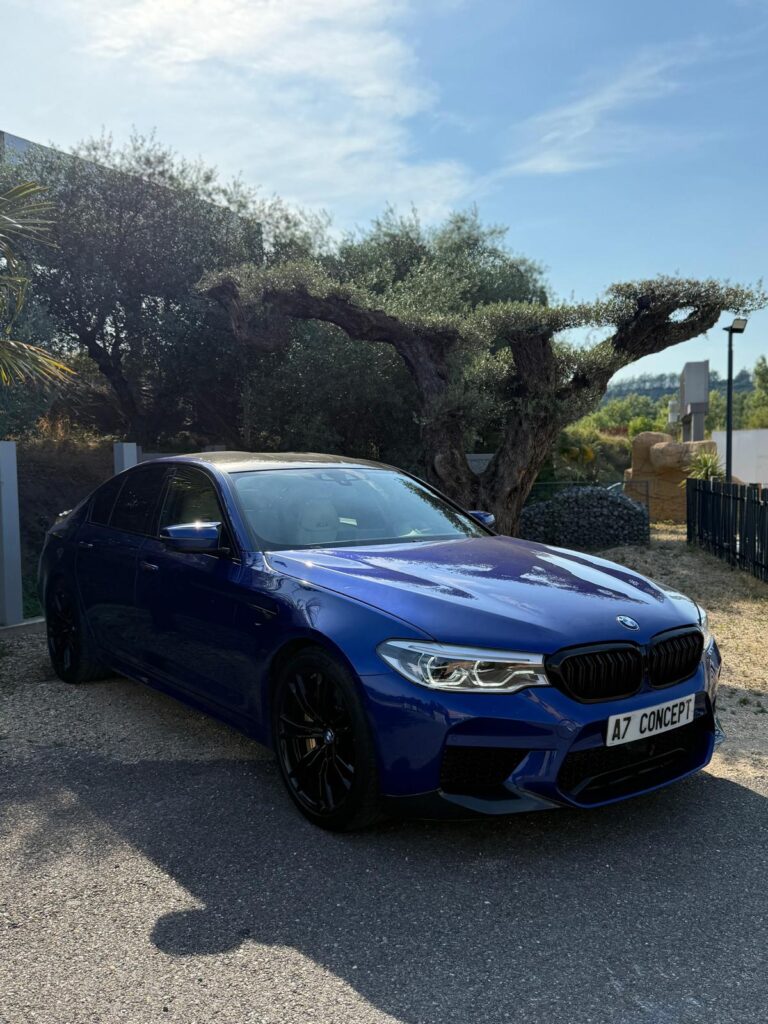 Superbe BMW M5 bleue nuit aux roues noires, garée sous un ciel bleu près d'un vieil olivier. Plaque "A7 CONCEPT".