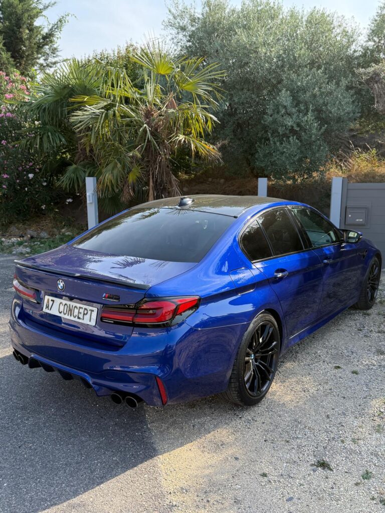 Arrière d'une BMW M5 Competition bleue électrique avec jantes noires, échappement quadruple et plaque 'A7 CONCEPT' dans un cadre ensoleillé.